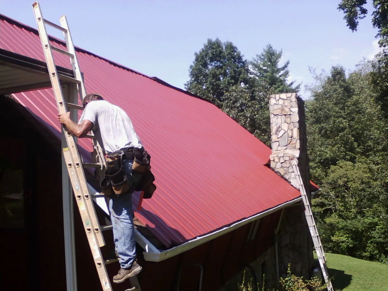 Roofer on a ladder beside a red metal roof and stone chimney — Soffit & Fascia Repair by Roofing Brothers in Spottsville